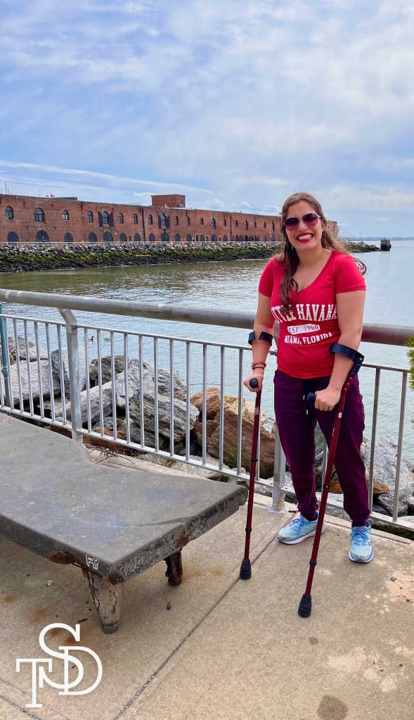 Me standing at a Waterfront with my forearm crutches, smiling and wearing sunglasses, a red shirt and purple pants with blue sneakers. There are loft style buildings and a body of water behind me.