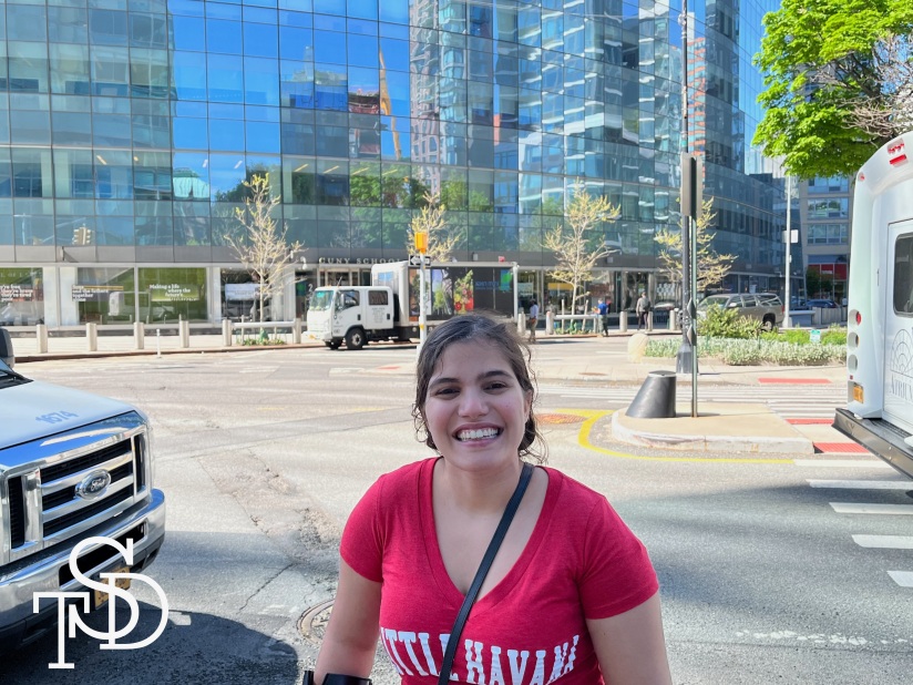 I'm standing in front of a busy intersection on a sunny day, wearing a red tee shirt and smiling. Behind me there's a big, tall glass building. A truck is parked in front of the main entrance. My logo is in the bottom corner in white.